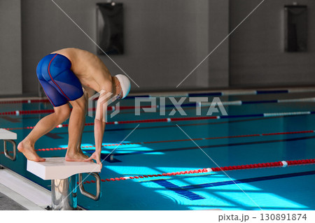 Swimmer bending on starting block before diving into pool for competitive race Swimmer bending on starting block before diving into pool for competitive race 130891874
