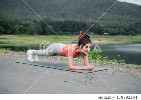 Woman in athletic wear doing a plank exercise on a yoga mat by a lake with green hills. Woman in athletic wear doing a plank exercise on a yoga mat by a lake with green hills. 130892141