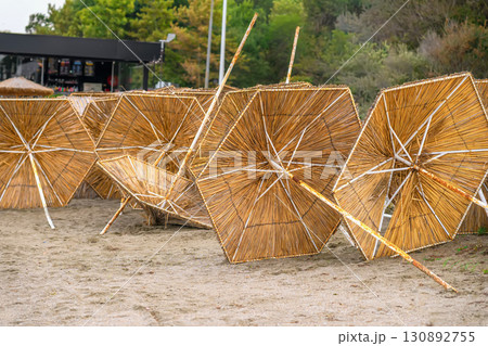 Straw Beach Umbrellas. A row of straw beach umbrellas lying on sandy ground near a beach. Concept of seaside relaxation, end of summer, end of holiday season Straw Beach Umbrellas. A row of straw beach umbrellas lying on sandy ground near a beach. Concept of seaside relaxation, end of summer, end of holiday season 130892755