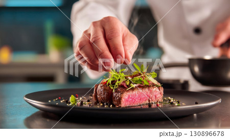 The chef decorates a medium-rare beef steak with fresh microgreens on a black plate, exquisite cuisine for gourmets 130896678