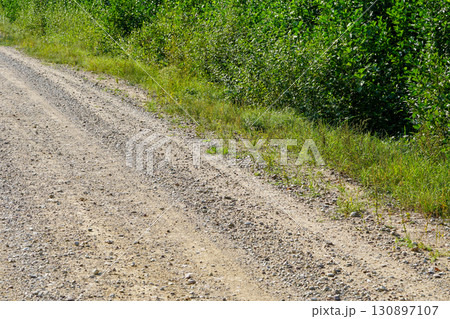 Rural gravel road with scattered stones and green vegetation on the roadside in summer sunlight Rural gravel road with scattered stones and green vegetation on the roadside in summer sunlight 130897107