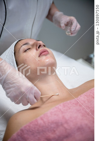 Beautician applying cosmetic treatment on female face with cotton swab in beauty salon 130897550