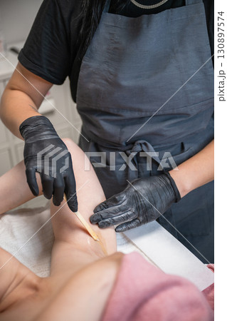 Beautician applying wax with wooden spatula on female armpit during professional hair removal procedure 130897574