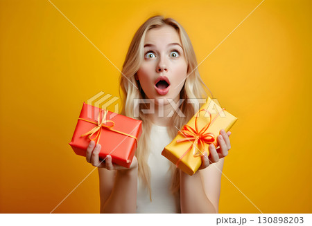 Surprise and excitement captured as young woman holds colorful gift boxes against a bright yellow backdrop 130898203