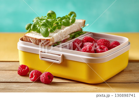 Yellow lunchbox with sandwich, greens and raspberries on wooden table. 130898741