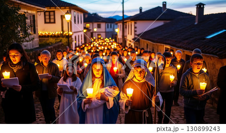 A group of people, including children, participate in a candlelit procession down a cobblestone street at dusk. They hold candles and wear costumes, illuminated by warm glowing lights 130899243