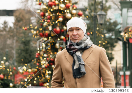 Man in cozy winter outfit stands outdoors near decorated Christmas tree, showcasing holiday decorations. Festive spirit of Xmas holiday with light garland. 130899548