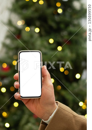 Man hand with smartphone with white blank screen against Christmas tree light garland and fairy lights. Festive Xmas Background. Mock up. Close up. 130899550