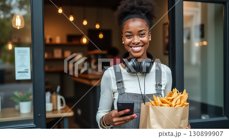 Smiling woman holding fries and smartphone outside a cozy eatery in the afternoon 130899907