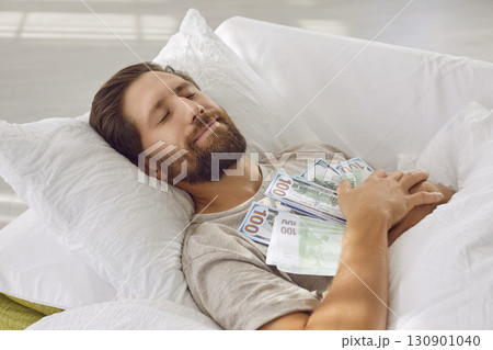 Smiling sleeping man lying in bed with closed eyes, embracing lots of cash dollar banknotes at home 130901040