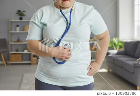 Fat woman standing at home and listening heartbeat on stethoscope after doing exercise. Fat woman standing at home and listening heartbeat on stethoscope after doing exercise. 130902380