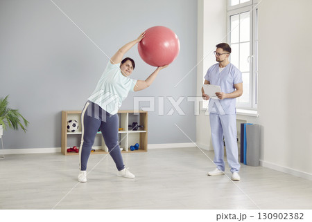 Overweight woman doing exercises with a fit ball under the control of a physiotherapist Overweight woman doing exercises with a fit ball under the control of a physiotherapist 130902382