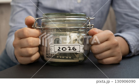 Unrecognizable woman holding Saving Money In Glass Jar filled with Dollars banknotes. 2026 year transcription in front of jar. Managing personal finances extra income for future insecurity 130906090