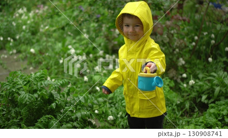 child in yellow raincoat with hood stands in the rain filling a small watering can with rainwater in front of a green garden 130908741