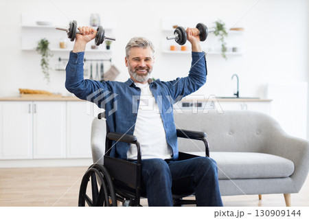 Mature Caucasian man using wheelchair lifting dumbbells to exercise at home living room 130909144