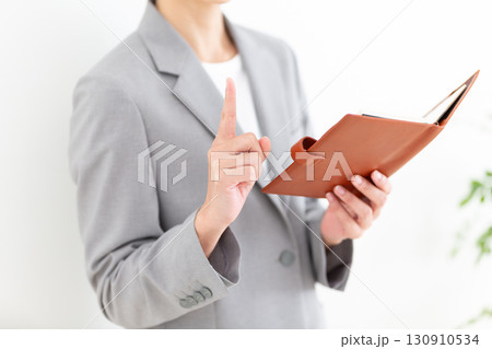 手帳を持って人差し指を立てるグレーのスーツ姿のミドル女性ビジネスウーマン 手帳を持って人差し指を立てるグレーのスーツ姿のミドル女性ビジネスウーマン 130910534
