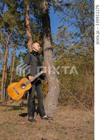 Portrait of smiling caucasian artist plays wooden acoustic guitar in autumnal park. Young songwriter plays string musical instrument hobby outside in nature fall time. Audio music healing Portrait of smiling caucasian artist plays wooden acoustic guitar in autumnal park. Young songwriter plays string musical instrument hobby outside in nature fall time. Audio music healing 130911276