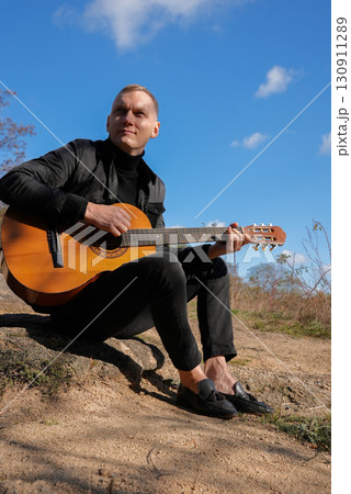 Beautiful blonde man playing acoustic guitar and singing relaxing enjoying outdoors on sunny autumn day. Artist learning and practicing playing musical instrument. Fall leaves autumnal 130911289