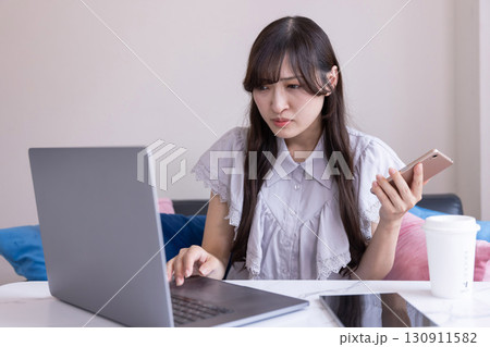 リビングで悩んだ表情を浮かべながらスマホを手にPC作業をする日本人女性 130911582