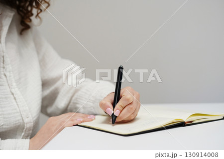 A woman in a white shirt is writing in a notebook with a black pen on a clean white desk A woman in a white shirt is writing in a notebook with a black pen on a clean white desk 130914008