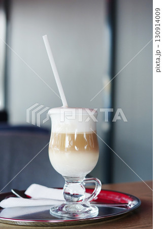 A layered latte with foamy milk served in a clear glass on a silver tray with a straw A layered latte with foamy milk served in a clear glass on a silver tray with a straw 130914009
