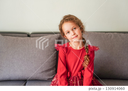 A young girl in a red festive dress sits on a gray couch, looking calmly at the camera 130914060