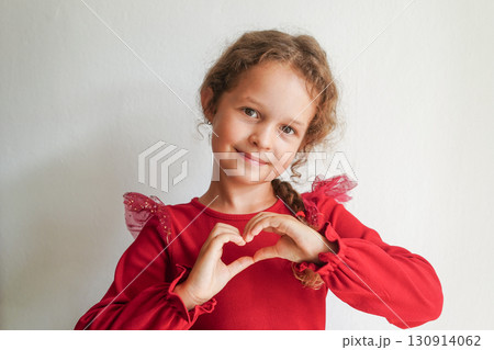 A smiling girl in a red dress forms a heart shape with her hands in front of a white wall 130914062