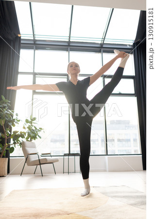 A young woman in sportswear practices a standing yoga pose indoors, raising one leg high and holding the foot with her hand for balance, strength, and flexibility in a bright modern living room A young woman in sportswear practices a standing yoga pose indoors, raising one leg high and holding the foot with her hand for balance, strength, and flexibility in a bright modern living room 130915181