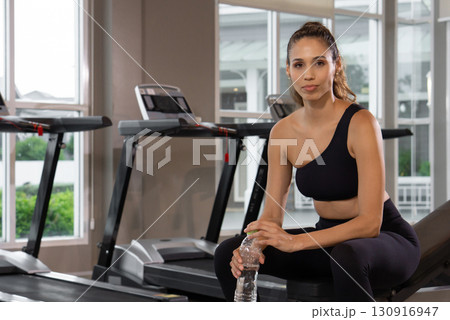 A fit individual take a break after exercising on a treadmill in a modern gym. The spacious setting has natural light and fitness equipment visible in the background. 130916947
