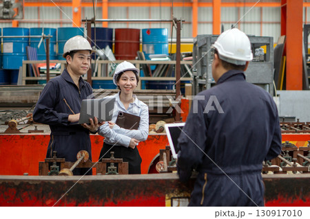 Three workers in a factory discuss a project using laptop computer. Everyone wearing protective hardhat. Atmosphere in manufactory with large machinery. Three workers in a factory discuss a project using laptop computer. Everyone wearing protective hardhat. Atmosphere in manufactory with large machinery. 130917010