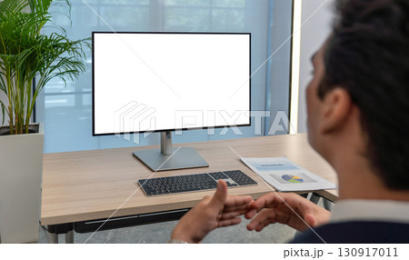 Individual is focused on a blank computer screen in a contemporary office environment. A document with a chart is visible on the desk, indicating preparation for a presentation. 130917011