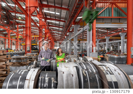 A woman in hardhat and safety vest operate industrial equipment inside a busy factory setting. Young male worker stand beside, check material while holding document clipboard. A woman in hardhat and safety vest operate industrial equipment inside a busy factory setting. Young male worker stand beside, check material while holding document clipboard. 130917179