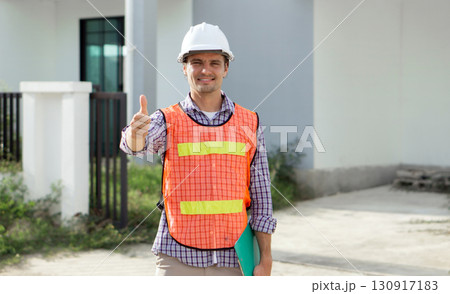 A construction worker wearing a hard hat and high-visibility vest stand at a building site. He smile and give a thumbs up, indicating a successful day of work. 130917183