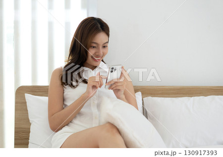 Young asian woman sit comfortably on a bed, engaging with her smartphone and smiling. The room has a minimalist design with soft pillow and bright lighting, creating a warm atmosphere. Young asian woman sit comfortably on a bed, engaging with her smartphone and smiling. The room has a minimalist design with soft pillow and bright lighting, creating a warm atmosphere. 130917393