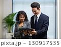 A man and woman dressed in business suit smile while examining a tablet in a contemporary office. Sunlight filters through large window, enhancing the productive atmosphere. 130917535