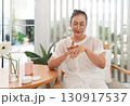 Senior asian woman sit at a wooden table, applying a skincare product to her hand. She enjoy bright natural light filtering through sheer curtains in a cozy home environment. 130917537