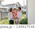 A family smile while standing in a developing neighborhood. The father hold their child in a safety vest and helmet while the mother stand beside them, both wearing casual clothing. 130917538