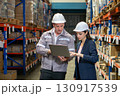 Business woman in suit and safety helmet review inventory record on laptop computer. Stand in a well-organized warehouse. Shelve are lined with various product, indicating a busy working environment 130917539