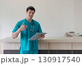 A young male healthcare worker in teal scrub stand at a reception desk in a well-lit clinic. He is holding a clipboard and looking confidently at the camera, ready for consultation. 130917540