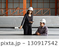 Two worker in hardhat talk in a warehouse. They discuss plans while standing near large, wrapped material. 130917542