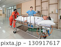 Two healthcare worker are hurriedly transferring a patient on a stretcher in a  well-lit hospital hallway during the day, illustrating urgency and teamwork in medical care. 130917617