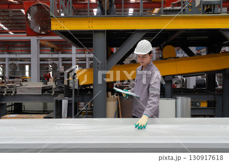 Young asian worker in safety helmet and protective gloves checking material covered in plastic. Work atmosphere in metal sheet manufactory. 130917618
