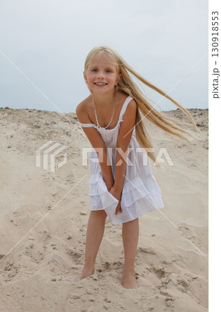 Joyful young blond child girl in white dress standing on sand outdoors 130918553