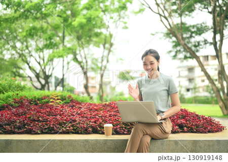 Young woman sitting on stone bench with laptop, waving during an online video call in a park 130919148
