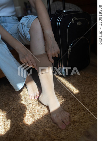 Woman putting on compression stockings before a long trip, suitcase in the background. Concept of leg protection during travel, varicose veins and deep vein thrombosis on long flights or car journeys 130919539