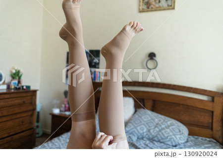 Woman putting on compression stockings in bedroom to prevent varicose veins. Swelling and leg pain, medical hosiery for circulation and vein health 130920024