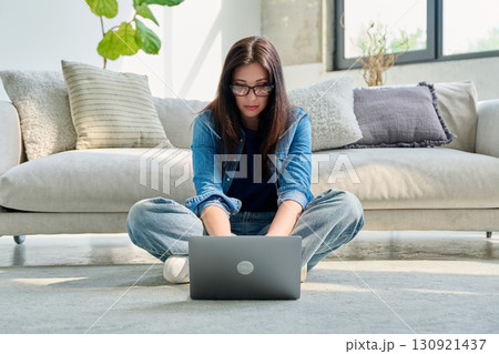 Young serious woman typing on laptop sitting on floor near couch at home 130921437