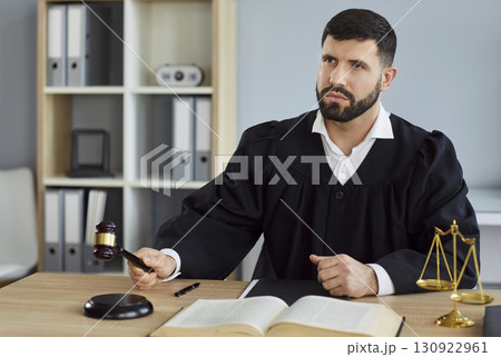 Male judge sitting on workplace in office in judicial robe knocking gavel having court verdict 130922961