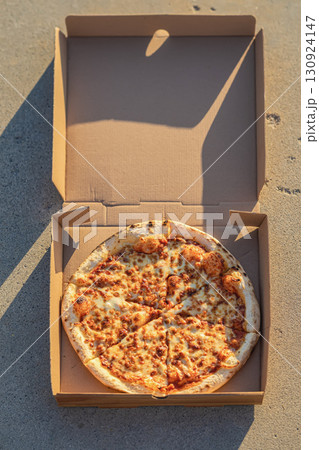 Freshly baked cheese pizza in an open cardboard box on sunny concrete background, melted cheese and golden crust. Top view. Concept of fast food craving, street food, casual outdoor meal Freshly baked cheese pizza in an open cardboard box on sunny concrete background, melted cheese and golden crust. Top view. Concept of fast food craving, street food, casual outdoor meal 130924147