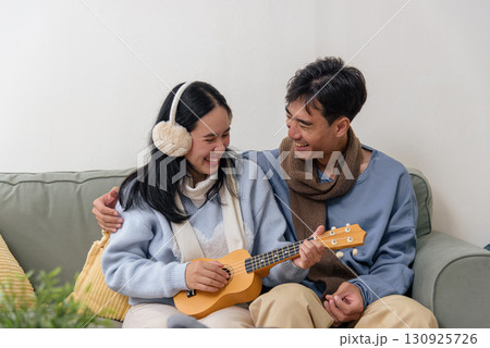 Winter day, happy couple enjoy family home indoors, people share loving moments, woman plays ukulele, man embraces her creating cozy warmth during cold winter season. 130925726
